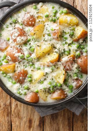 Potatoes and green peas baked in creamy cheese sauce close-up in a frying pan on a wooden table. Vertical top view 112925604