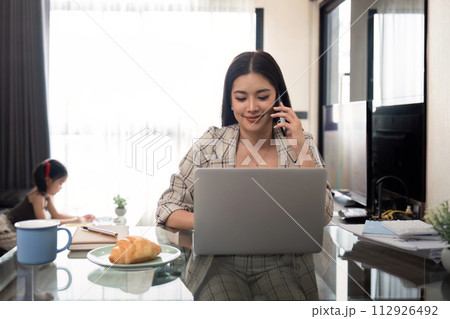 Asian female speaking on mobile phone while using laptop for remote work taking care of daughter at home 112926492