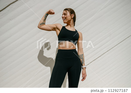 Smiling female model in sportswear showing biceps standing outdoors and looks away Smiling female model in sportswear showing biceps standing outdoors and looks away 112927187