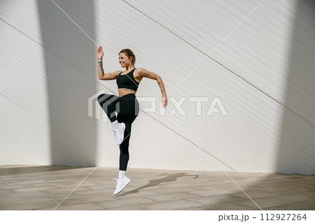 Female athlete in sportswear leaping in air with wall backdrop outdoors. Healthy lifestyle concept 112927264