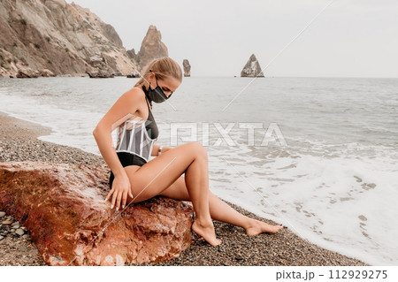 Woman travel portrait. Happy woman with long hair poses on a red volcanic rock at the beach. Close up portrait cute woman in black bikini, smiles at the camera, with the sea in the background. 112929275