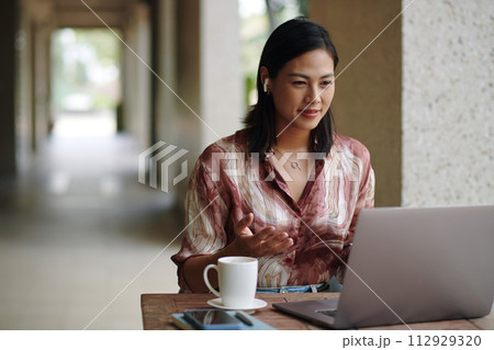 Smiling businesswoman sitting at coffeeshop table and speaking to colleagues in online meeting Smiling businesswoman sitting at coffeeshop table and speaking to colleagues in online meeting 112929320