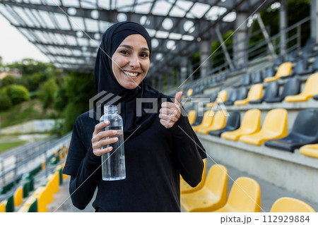 Active woman in a hijab holding a water bottle, giving a thumbs up at an outdoor stadium, showing hydration and health. Active woman in a hijab holding a water bottle, giving a thumbs up at an outdoor stadium, showing hydration and health. 112929884