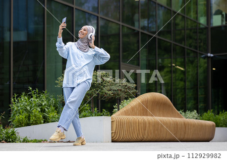 A young, fashionable woman wearing a hijab captures a selfie outdoors with greenery and modern architecture in the backdrop. A young, fashionable woman wearing a hijab captures a selfie outdoors with greenery and modern architecture in the backdrop. 112929982