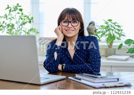 Middle aged woman sitting at workplace in home office, holding pet parrot on shoulder 112931239