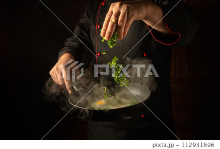 Cropped silhouette of a chef with a hot frying pan for cooking scrambled eggs for breakfast. The cook hand tosses fresh parsley into fried eggs for aroma and taste 112931696