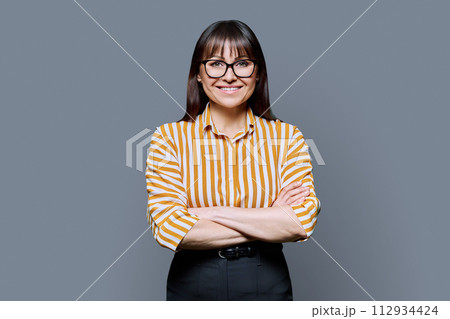 Portrait of confident smiling middle aged woman with crossed arms on grey background Portrait of confident smiling middle aged woman with crossed arms on grey background 112934424
