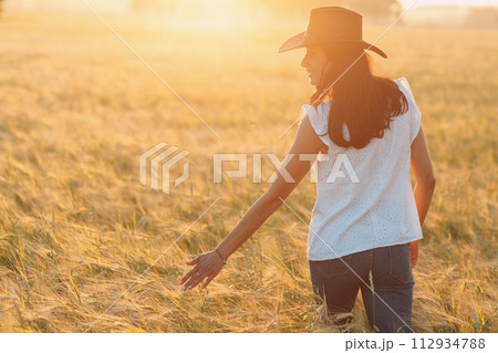 Woman farmer in cowboy hat walking with hands on ears at agricultural barley field on sunset. 112934788