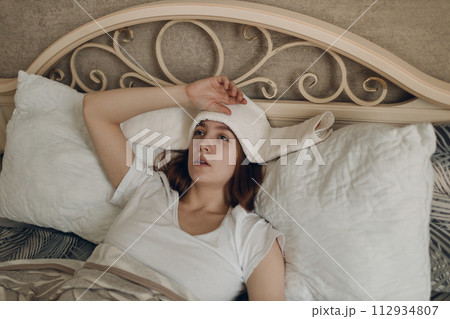 Young woman with headache flu ill sick disease cold at home indoor lying on bed with cold compress on her head at bedroom home Young woman with headache flu ill sick disease cold at home indoor lying on bed with cold compress on her head at bedroom home 112934807