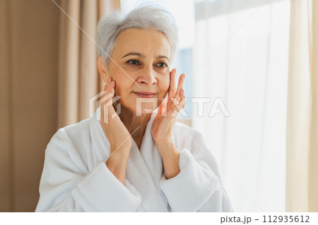 Mature adult old woman applying nourishing anti age face cream doing daily beauty routine. Senior woman touching face skin wrinkles gets facial treatment. Skin care aging beauty skincare cosmetics Mature adult old woman applying nourishing anti age face cream doing daily beauty routine. Senior woman touching face skin wrinkles gets facial treatment. Skin care aging beauty skincare cosmetics 112935612