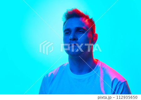 Portrait of handsome young man in white t-shirt looking away confidently in neon light against gradient blue background. Portrait of handsome young man in white t-shirt looking away confidently in neon light against gradient blue background. 112935650