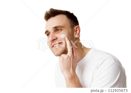 Young handsome man applying facial care, day cream and looking away against white studio background. 112935673