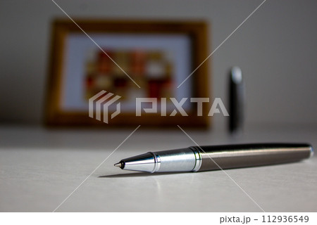 Pen on beautiful white fon on warm sunlight. Wooden picture frame and cap in the background. Pen on beautiful white fon on warm sunlight. Wooden picture frame and cap in the background. 112936549