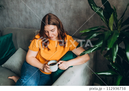 Young woman wears yellow t-shirt drink tea or coffee and sit on the sofa couch at home, hotel, or flat. 30s female rest and spend free spare time in living room indoors. People lounge concept Young woman wears yellow t-shirt drink tea or coffee and sit on the sofa couch at home, hotel, or flat. 30s female rest and spend free spare time in living room indoors. People lounge concept 112936738