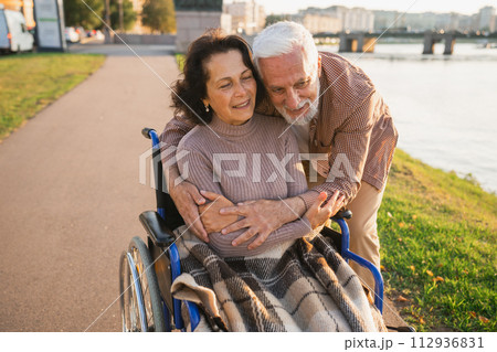 Senior woman in wheelchair walking with caregiver old man on road in park. Elderly family couple man supporting embracing woman in chair for people with disability outdoor. Rehabilitation 112936831