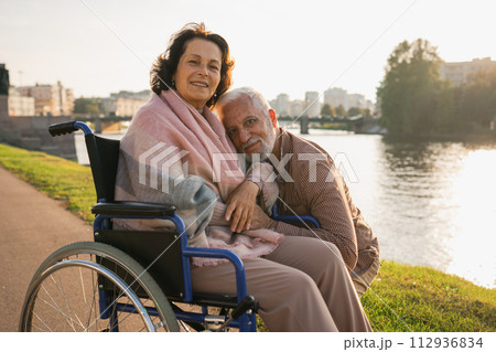 Senior woman in wheelchair walking with caregiver old man on road. Elderly family couple man supporting taking care of paralyzed woman in chair for people with disability outdoor. Rehabilitation 112936834