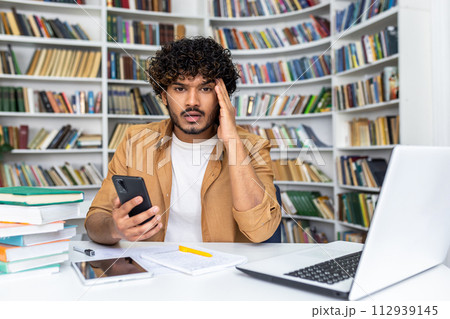 Overwhelmed islamic man sitting at desk and holding portable smartphone in personal cabinet with designer bookshelves on background. Workplace with wireless laptop, digital tablet and papers. 112939145
