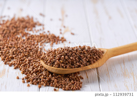 Raw buckwheat crumble from in a wooden spoon on a white wooden table. Raw buckwheat crumble from in a wooden spoon on a white wooden table. 112939731