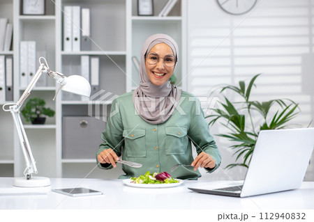 Positive muslim woman sitting at workplace and having break with salad while working on wireless laptop. Office equipped with adjustable lamp and various supplies organized on shelves on background. 112940832