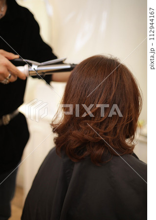 女性の髪にヘアアイロンをかける男性美容師 112944167