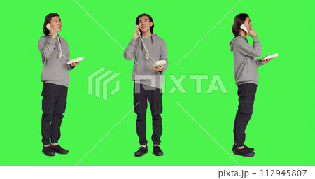 Asian guy talking on landline phone, answering call on telephone with cord while he stands against full body greenscreen. Young person using retro office phone for remote communication. 112945807