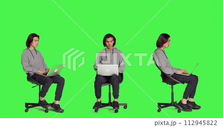 Young adult browsing the web on laptop in full body greenscreen studio, using mobile computer to navigate through webpages online. Person sitting on chair and working on wireless pc, trendy guy. Young adult browsing the web on laptop in full body greenscreen studio, using mobile computer to navigate through webpages online. Person sitting on chair and working on wireless pc, trendy guy. 112945822