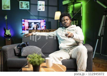 Man zapping through TV broadcast channels using remote, relaxing in living room used as home theatre. BIPOC person in RGB lit apartment sitting on sofa, using television set to have fun 112945872