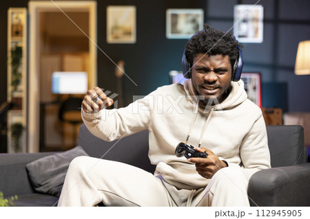 BIPOC player shouting after losing videogame, doing frustration gestures. Angry gamer on couch in apartment ready to throw gamepad controller, raging after receiving game over message BIPOC player shouting after losing videogame, doing frustration gestures. Angry gamer on couch in apartment ready to throw gamepad controller, raging after receiving game over message 112945905