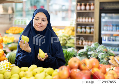 Happy girl in paranja making purchases in supermarket, choosing fresh ripe pears 112947055