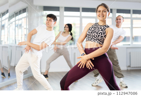 Happy girl and group of young active sport people practicing vogue dancing movements while training twist or charleston in dance hall 112947086