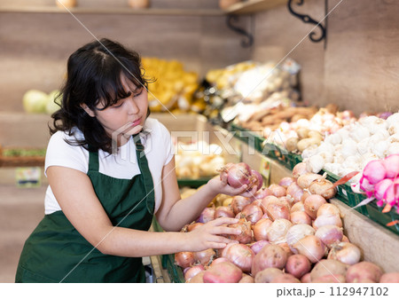 Attentive young saleswoman placing onions on food stall in grocery store Attentive young saleswoman placing onions on food stall in grocery store 112947102