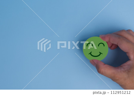 customer service evaluation and satisfaction survey concept. hand holding emotion happy smile face icon on wooden cube on blue background. copy space 112951212