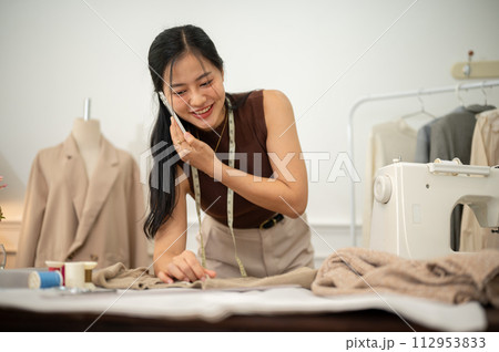 A professional, busy female tailor is talking on the phone with while working in her studio. A professional, busy female tailor is talking on the phone with while working in her studio. 112953833
