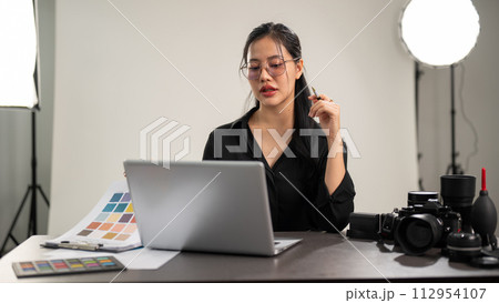 A confident Asian female photographer is working on her laptop, working in a photoshoot studio. A confident Asian female photographer is working on her laptop, working in a photoshoot studio. 112954107