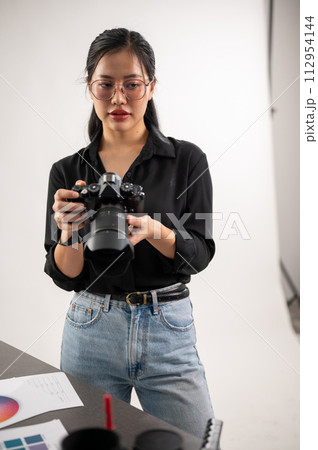 A confident, experienced Asian female photographer is adjusting her DSLR camera, preparing photoshoot equipment, and working in the studio. A confident, experienced Asian female photographer is adjusting her DSLR camera, preparing photoshoot equipment, and working in the studio. 112954144