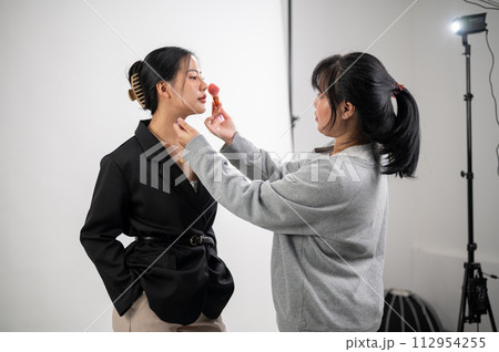 A female model is having makeup applied by a makeup artist, preparing for a photoshoot in the studio A female model is having makeup applied by a makeup artist, preparing for a photoshoot in the studio 112954255