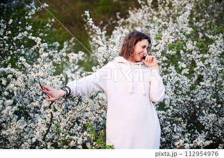 Woman allergic suffering from seasonal allergy at spring in blossoming garden at springtime. Young woman sneezing, closing nose by hand in front of blooming tree. Spring allergy concept 112954976