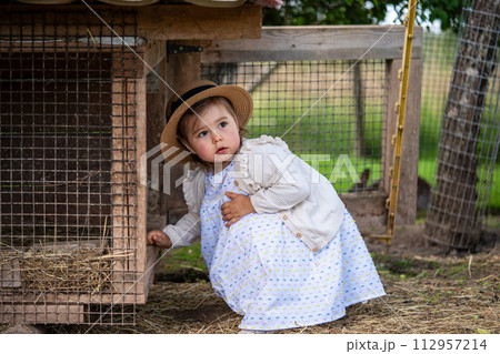 a cute little girl in a dress and hat sits near a rabbit house on a farm  112957214