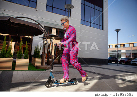 Stylishly dressed mature woman in a bright pink pantsuit of trousers and a jacket rides around the city on a scooter, an active, healthy and eco friendly lifestyle Stylishly dressed mature woman in a bright pink pantsuit of trousers and a jacket rides around the city on a scooter, an active, healthy and eco friendly lifestyle 112958583
