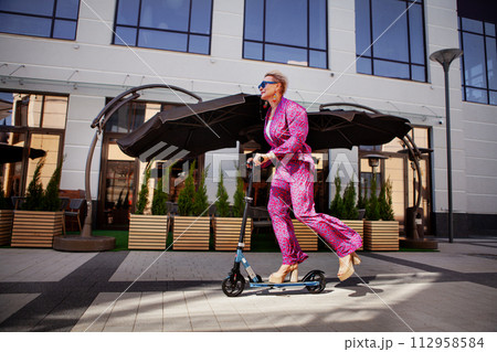 Stylishly dressed mature woman in a bright pink pantsuit of trousers and a jacket rides around the city on a scooter, an active, healthy and eco friendly lifestyle Stylishly dressed mature woman in a bright pink pantsuit of trousers and a jacket rides around the city on a scooter, an active, healthy and eco friendly lifestyle 112958584