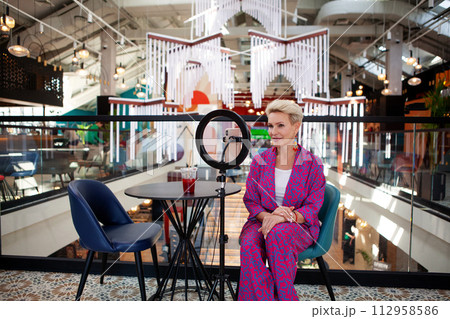 Beautiful mature blonde woman with a short haircut, an influencer, records a video blog while sitting at a table in a restaurant. Middle aged female blogger Beautiful mature blonde woman with a short haircut, an influencer, records a video blog while sitting at a table in a restaurant. Middle aged female blogger 112958586