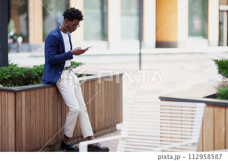 Stylish African American man in smart casual clothes, with mobile phone against backdrop of city. Fashionable guy with smartphone outdoors Stylish African American man in smart casual clothes, with mobile phone against backdrop of city. Fashionable guy with smartphone outdoors 112958587