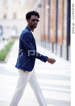 Handsome fashion African American man walks down the street in stylish attire white pants, blue blazer. Fashionable street style trend for men Handsome fashion African American man walks down the street in stylish attire white pants, blue blazer. Fashionable street style trend for men 112958588