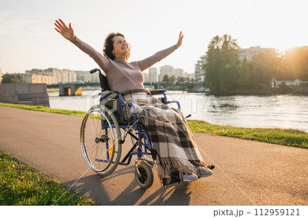 Happy senior woman in wheelchair raised hands enjoying freedom walking on road in city park outdoor. Old happy woman in chair for people with disability has a hope. Disability disease rehabilitation 112959121