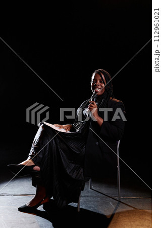 Vertical full length portrait of smiling young woman speaking to microphone on stage while sitting in chair with spotlight 112961021
