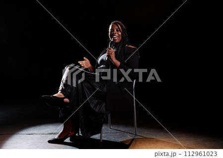 Minimal full length portrait of smiling Black woman performing comedy show while sitting in chair on stage and speaking to microphone copy space 112961023