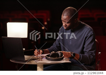 Portrait of Black adult man as theater director taking notes sitting at table with laptop copy space 112961044