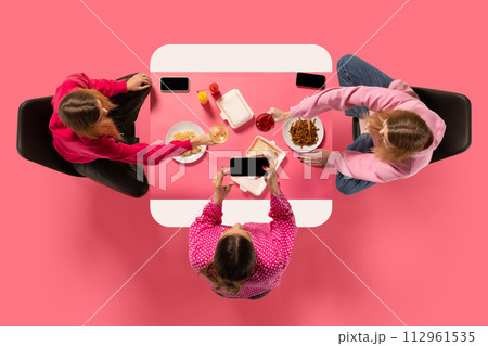 Creative collage. Top view portrait of two young girl sitting at table eating and speaking against pink background. Creative collage. Top view portrait of two young girl sitting at table eating and speaking against pink background. 112961535