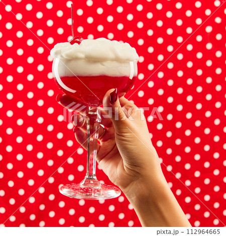 Sweet and sour. Hand holding cocktail glass decorated with cherry against red background with white polka dot. 112964665