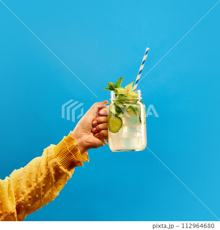 Female hand holding glass of refreshing cold citrus lemonade, mojito with straw against blue background. 112964680